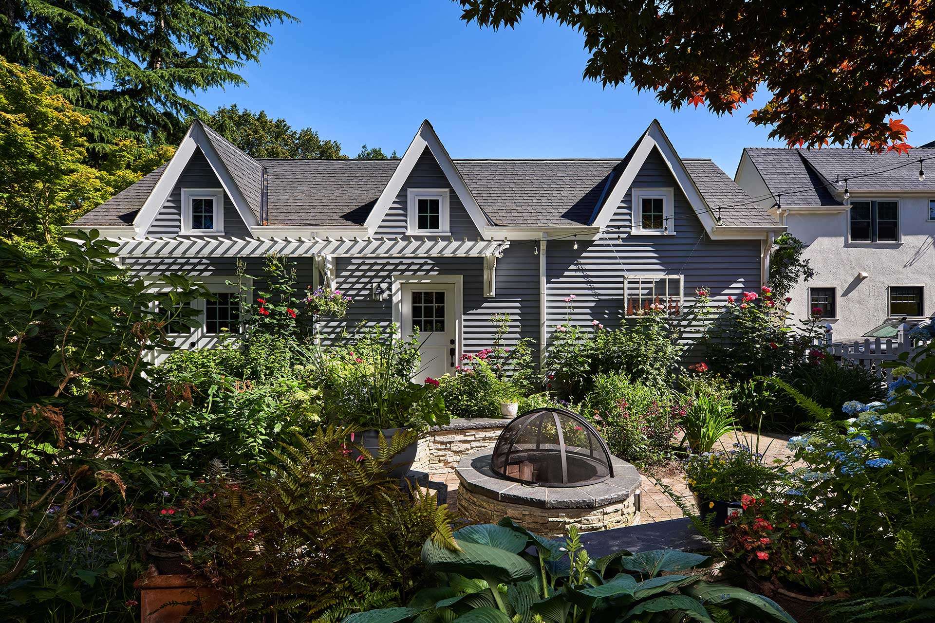 Three tall roof gables are featured on the side of the new garage design. This side of the garage faces the backyard, which is filled with plants and a fire pit.