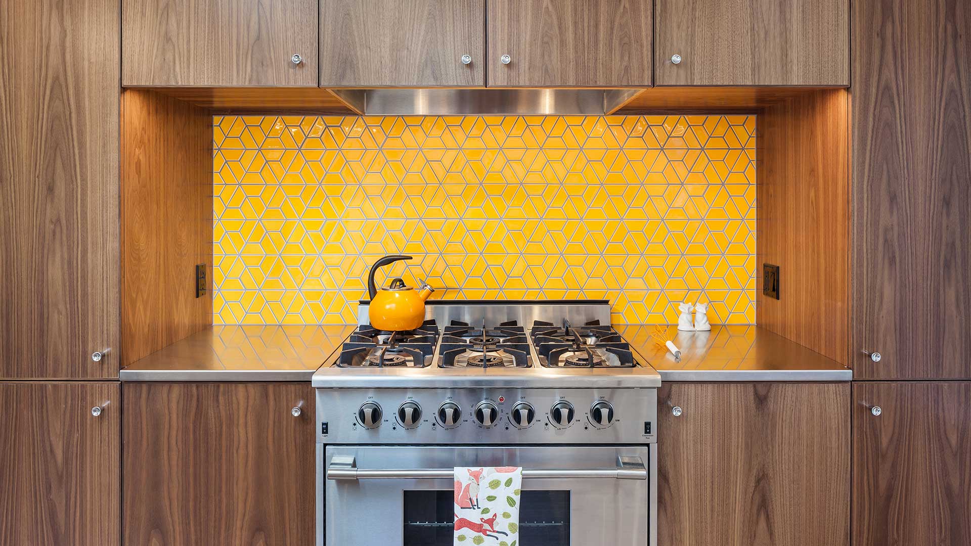A bright orange-colored tile backsplash sits behind the stainless steel gas range at the Hawkridge Modern. The surrounding cabinets are plain-sliced walnut. The countertop is stainless steel.