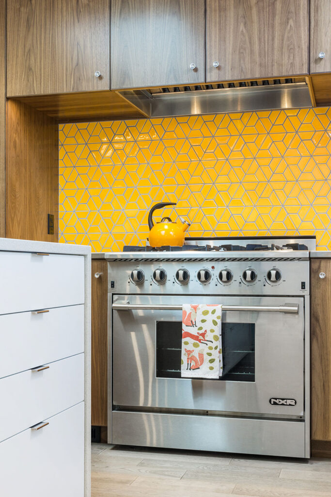 An orange-colored backsplash made of ceramic tile sits behind the stainless steel gas range after the residential remodel. The cabinetry is plain-sliced walnut.
