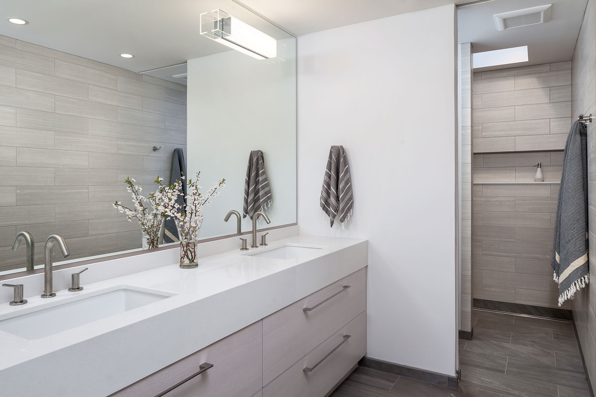 A large mirror above the sink vanity creates the illusion of a larger space in the primary bathroom. A cureless, walk-in shower with a tiled niche is in the background.