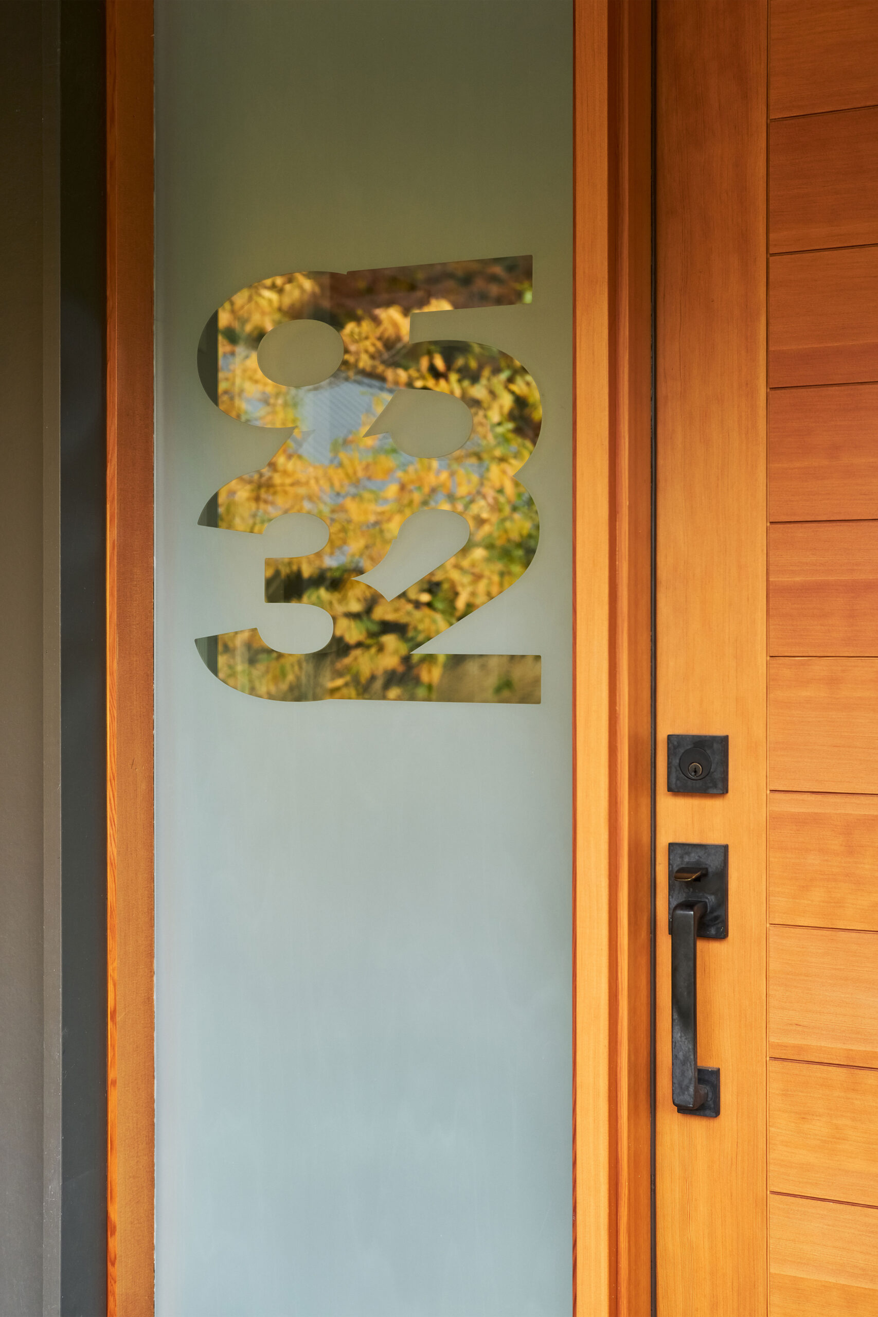 A glass sidelight features the street address numbers 9 5 3 2. The adjacent door is made of fir and features horizontally jointed wood and black hardware.