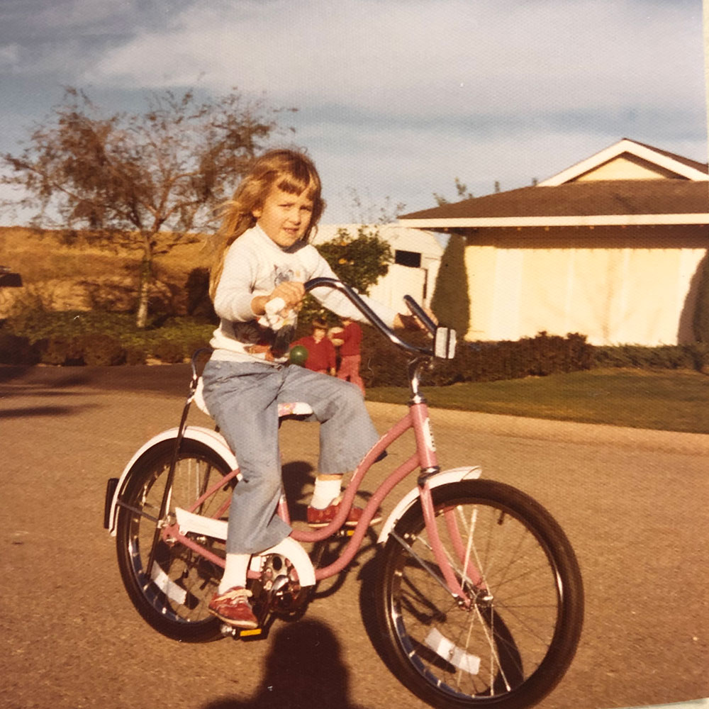 Young girl riding a pink bicycle in a suburban neighborhood, with a house and trees in the background.