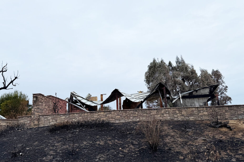 Collapsed metal roof draped over charred supports and stone walls after the Mountain Fire, showing how building materials respond differently to wildfire.
