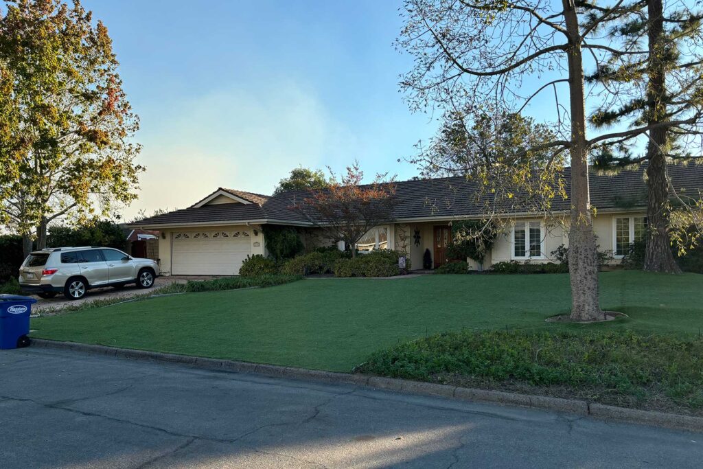 A single-story suburban house with a dark metal roof and artificial turf lawn, showing how wildfire resistant home design details can blend into an attractive residential setting.