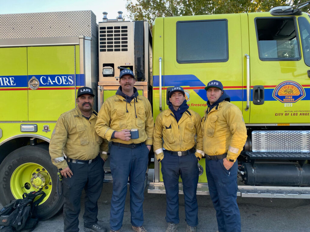 Four firefighters in yellow jackets standing in front of their fire engine, part of the crew who helped protect homes during the Mountain Fire in Camarillo.