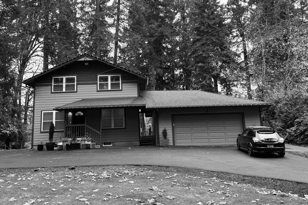 Existing 1970s house with a simple box form and an attached garage set lower than the main floor, connected by a partial breezeway and featuring minimal exterior detail.