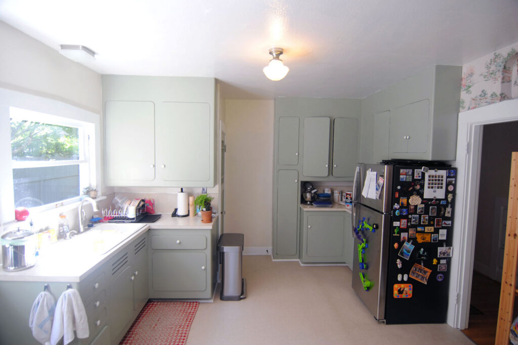 Existing kitchen before renovation showing painted cabinets, a freestanding refrigerator, minimal counter space, and multiple doorways breaking up the layout.