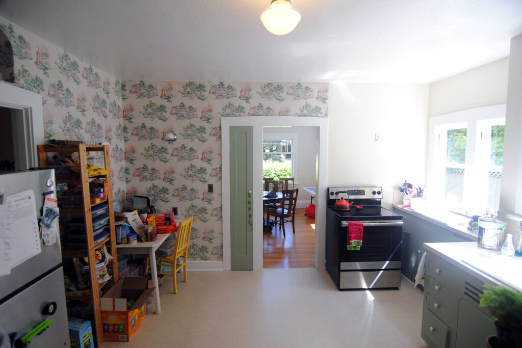Existing kitchen before renovation with wallpapered walls, a standalone range near a window, limited counter space, open shelving, and a doorway creating circulation through the room.