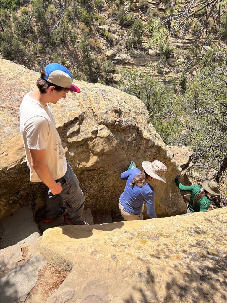 Visitors navigating a tight, carved rock passage with steps and handholds along the approach to a cliff dwelling.