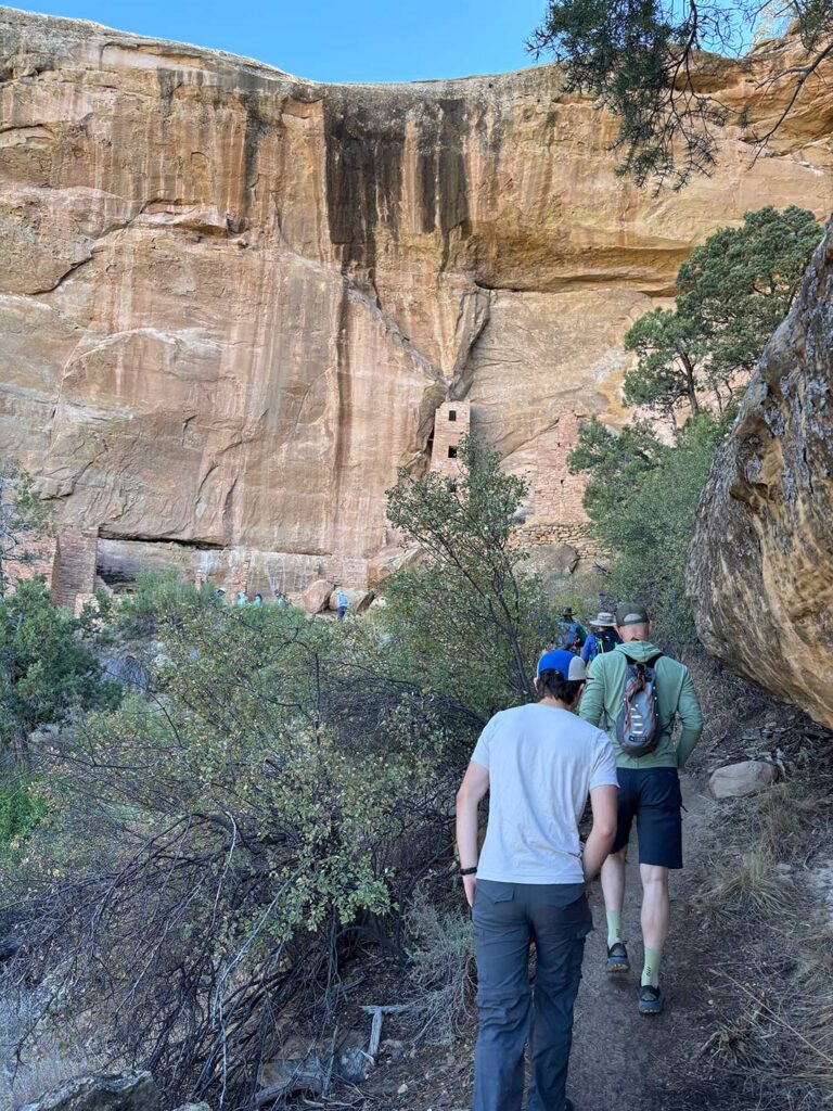 First view of a cliff dwelling tucked into a canyon wall at Mesa Verde National Park, with visitors approaching along a narrow trail.