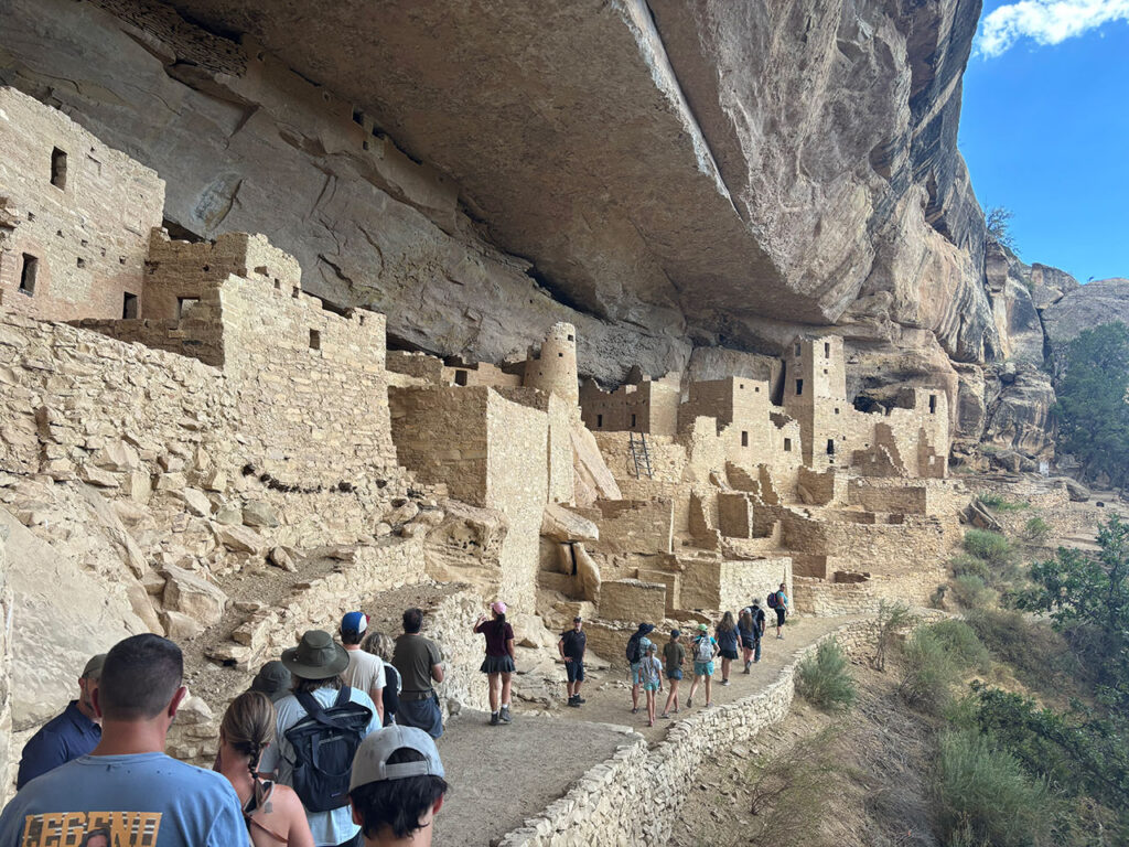 Visitors walking along a narrow path beside cliff dwellings at Mesa Verde National Park, with stone walls and rooms rising along the rock face.