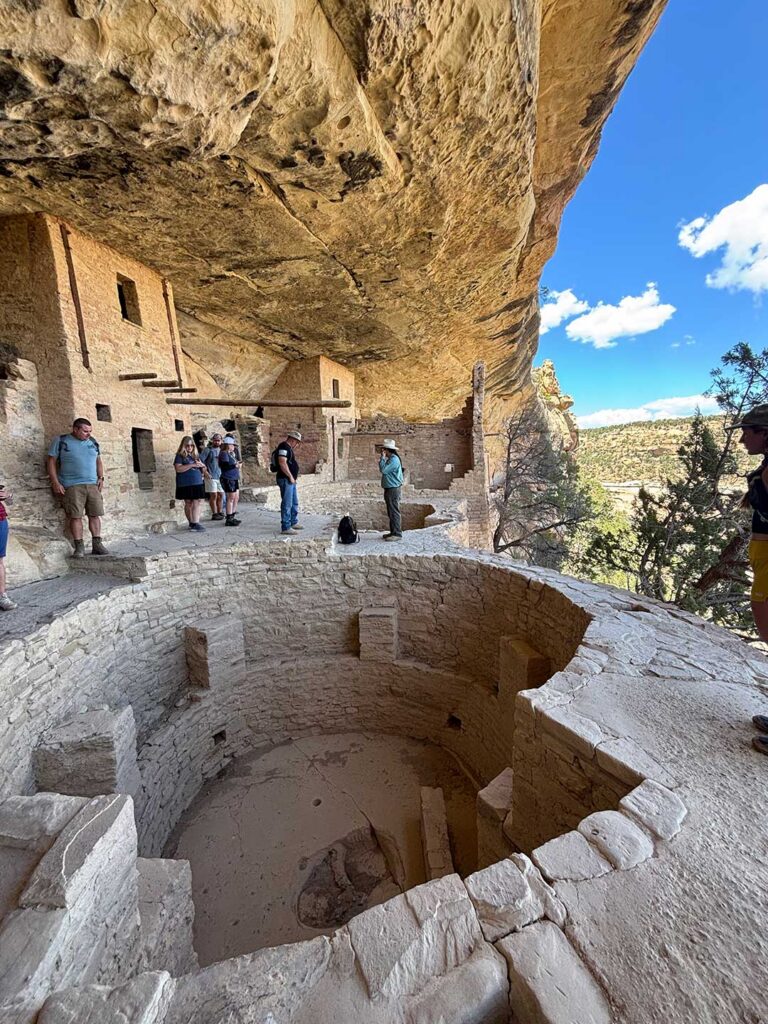Visitors standing at the edge of a circular kiva within a cliff dwelling at Mesa Verde National Park, showing the sunken form and surrounding stone structures.