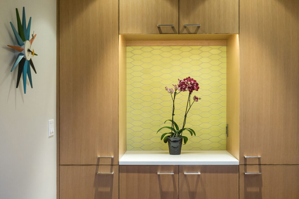 Frameless overlay cabinetry in natural wood with flat-panel doors and drawers, featuring an integrated niche with a yellow geometric tile backsplash and a potted orchid.