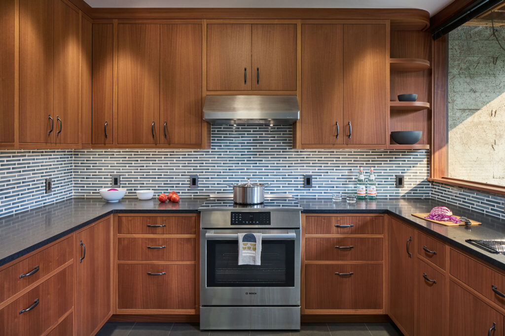 Natural mahogany flush inset cabinetry with slab doors and drawers, paired with a black granite countertop, stainless steel range and hood, and blue rectangular tile backsplash in a modern basement kitchen.