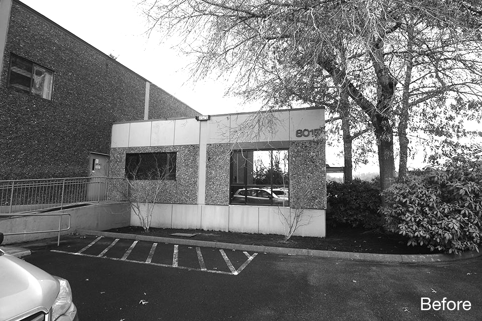Black-and-white photo of an existing one-story commercial building with pebble-dash exterior, small windows, and a narrow entry, set beside a parking area and mature trees.