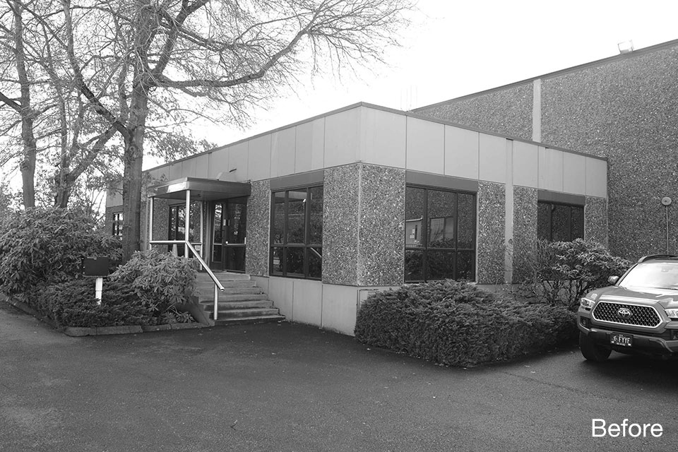Before photo of commercial building corner entry with small canopy, steps and railing, dated façade materials, and surrounding overgrown landscaping.