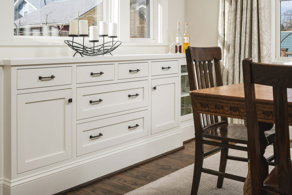 Flush inset built-in cabinetry with painted shaker-style doors and drawers, black hardware, and a painted wood countertop, located in a dining room with windows above and a wood dining table and chairs adjacent.