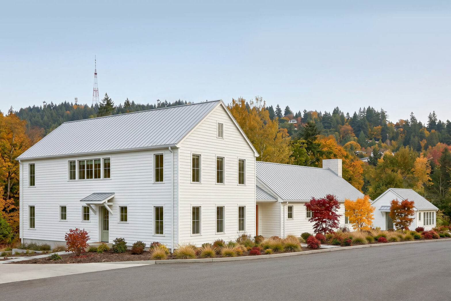 Oblique view of a new modern farmhouse with white siding and a standing seam metal roof, showing the main gabled volume and connected wings stepping down along a landscaped street with fall trees in the background.