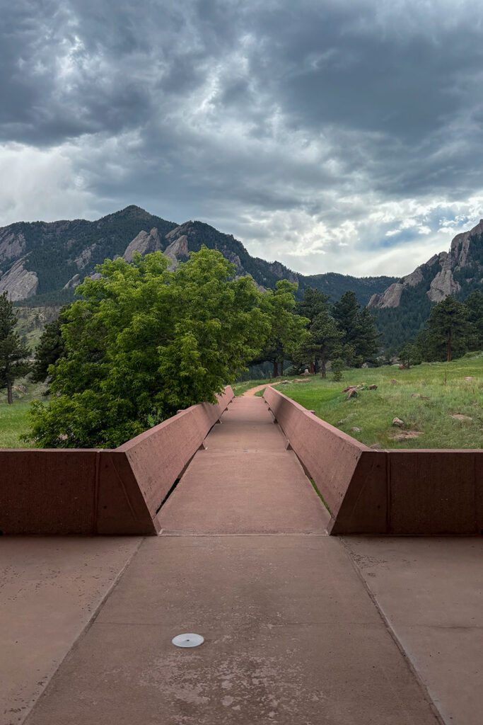 Concrete bridge extending into an open meadow, with the Flatirons, pine trees, and dramatic storm clouds beyond.