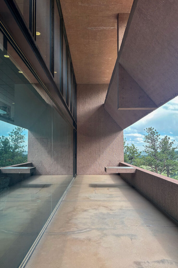Exterior walkway with reflective glass, exposed concrete walls and ceiling, a cantilevered concrete table, and views of pine trees beyond.